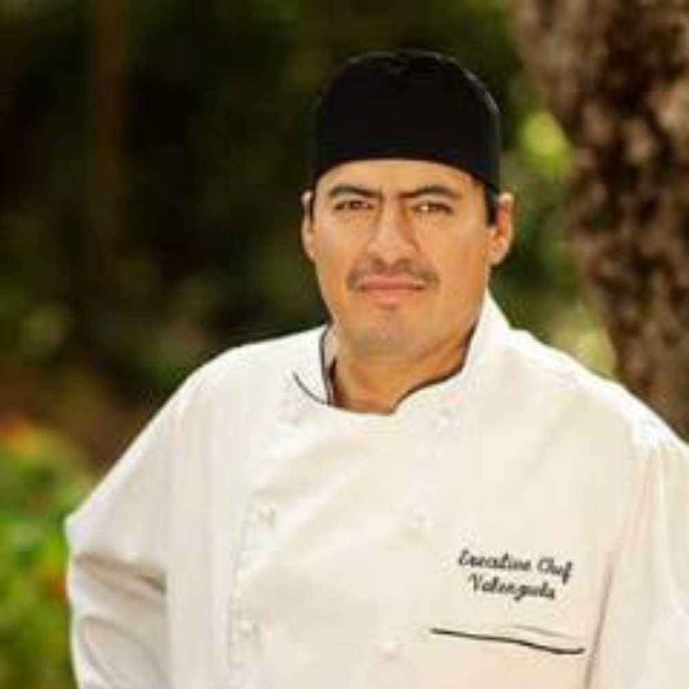 A man wearing a chef's uniform and black chef's hat stands outdoors with trees in the background. He has a serious expression and his hands are on his hips. The uniform reads "Executive Chef Velazques".